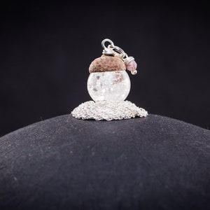 Puede incluir: Un collar de cadena de plata con un colgante de esfera de vidrio transparente. La esfera está coronada con una bellota marrón y una pequeña piedra rosa.