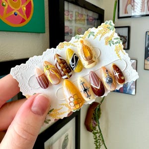 May include: A set of ten artificial nails in various designs and colors. The nails feature patterns including floral, geometric, and abstract art. Colors include brown, yellow, white, and orange. The nails are held on a decorative card.