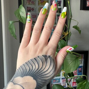 May include: A hand with a black and white tattoo is shown with green and white nail polish. The nails are decorated with colorful designs, including flowers, dots, and a heart.