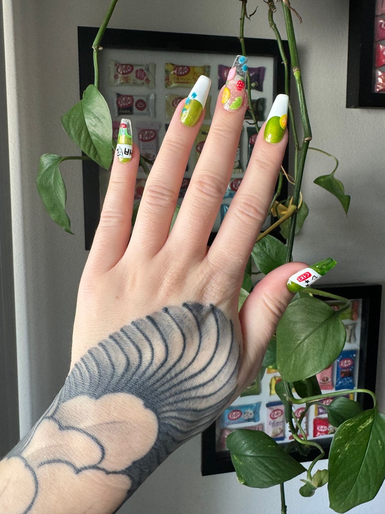 May include: A hand with a black and white tattoo is shown with green and white nail polish. The nails are decorated with colorful designs, including flowers, dots, and a heart.