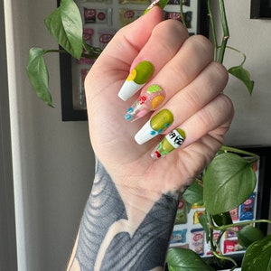 May include: Close-up of a hand with long, white, and clear acrylic nails. The nails are decorated with green, yellow, and red fruit designs. The nails are painted with a white base and have a clear top coat. The hand is wearing a black tattoo.