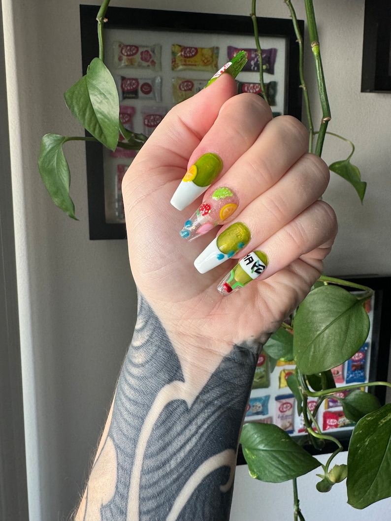 May include: Close-up of a hand with long, white, and clear acrylic nails. The nails are decorated with green, yellow, and red fruit designs. The nails are painted with a white base and have a clear top coat. The hand is wearing a black tattoo.