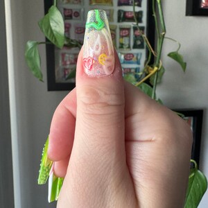 May include: A long, clear, square-shaped nail with a green, yellow, and red abstract design. The nail is decorated with glitter and small white dots.