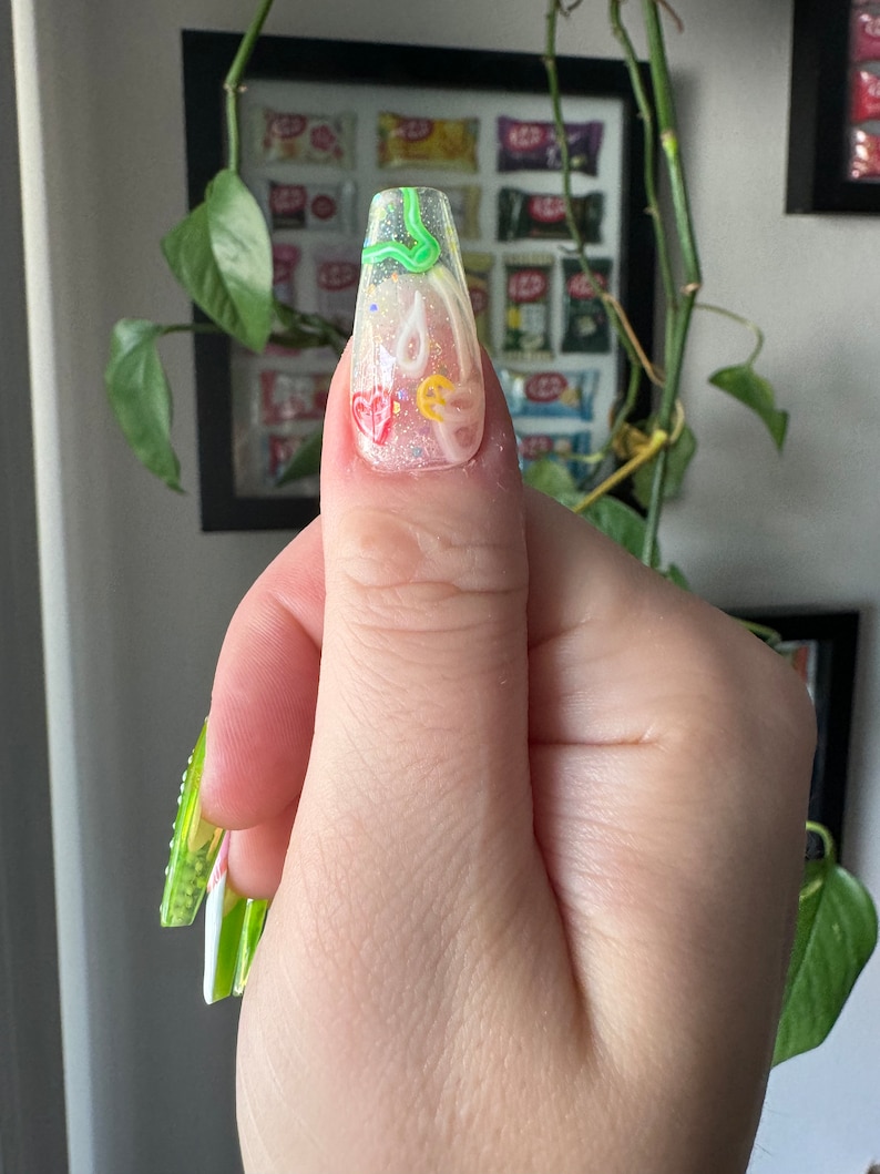 May include: A long, clear, square-shaped nail with a green, yellow, and red abstract design. The nail is decorated with glitter and small white dots.
