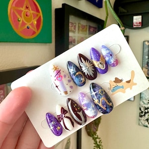 May include: A set of ten artificial nails with diverse designs. The nails feature glitter, celestial patterns, bows, and a crown. Colours include purple, white, brown, and blue. The nails are displayed on a white card.