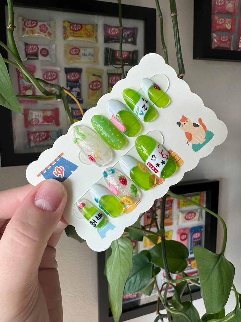 May include: A set of ten green and white press-on nails with various designs, including a strawberry, a lemon, a bottle of green juice, and a cat. The nails are displayed on a white card with a scalloped edge.