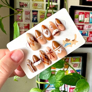 May include: Set of ten brown and beige false nails with different designs. The designs include a heart, a bow, and a plaid pattern.