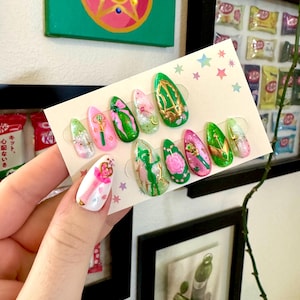 May include: A set of ten artificial nails with various designs. The nails feature a mix of pink, green, and white colors, with gold accents and floral patterns. Some nails have a Sailor Moon theme, with a star and moon design.