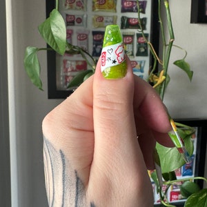 May include: A lime green nail with a white sticker featuring a cartoon character, two stars, and a heart. The sticker has the text "CIKI" on it.