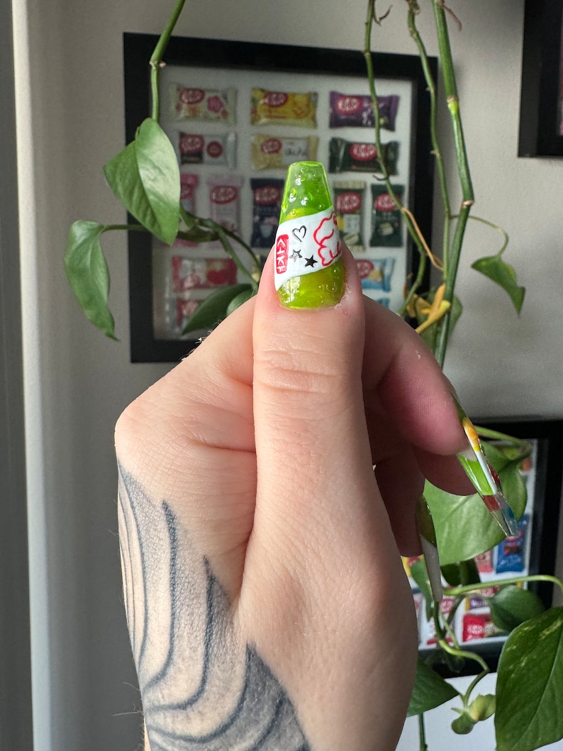 May include: A lime green nail with a white sticker featuring a cartoon character, two stars, and a heart. The sticker has the text "CIKI" on it.