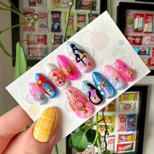 Può includere: Un set di dieci unghie finte a tema Sailor Moon. Le unghie sono dipinte in varie tonalità di rosa, blu e bianco con accenti di glitter e oro. Ogni unghia presenta un diverso personaggio o simbolo di Sailor Moon, tra cui Sailor Moon, Luna e lo scettro lunare.