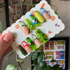 May include: A set of ten false nails with a green and yellow cucumber design. The nails are displayed on a white card with a citrus fruit design.