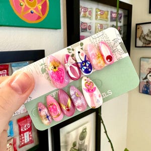 Può includere: Un set di unghie finte decorate a forma di mandorla. Le unghie presentano vari disegni, tra cui arte a tema Sailor Moon, abbellimenti dorati e schemi di colori rosa e bianco. Le unghie sono esposte su una carta verde.