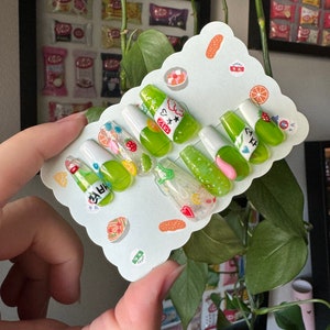 May include: A set of ten green and white press-on nails with a green soda bottle design. The nails have a variety of designs, including hearts, stars, and glitter. The nails are displayed on a white card with a scalloped edge.