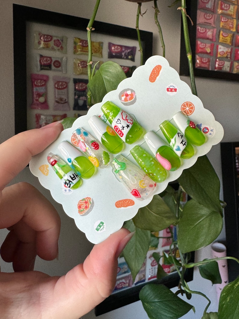 May include: A set of ten green and white press-on nails with a green soda bottle design. The nails have a variety of designs, including hearts, stars, and glitter. The nails are displayed on a white card with a scalloped edge.