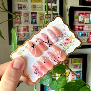 May include: A set of pink and white press-on nails with various designs, including bows, glitter, and a checkered pattern. The nails are displayed on a white card with a floral design.