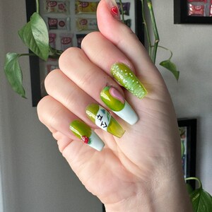 May include: Green and white long false nails with a green avocado design and the text "avocado" on one nail.  Another nail has a small red strawberry design.