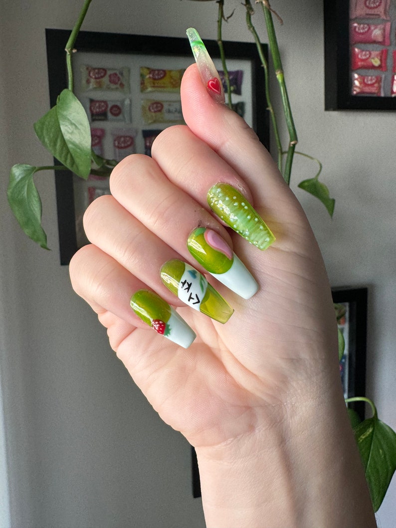 May include: Green and white long false nails with a green avocado design and the text "avocado" on one nail.  Another nail has a small red strawberry design.