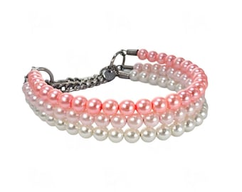 May include: A multi-strand bracelet featuring three rows of round beads in graduated shades of pink, light pink, and white. The strands are connected by dark metal hardware, including circular rings and a chain link section at the closure.