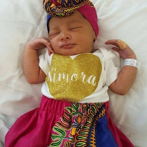 May include: A newborn baby wearing a white onesie with the name "Kimora" in gold glitter, a pink skirt with a colorful African print, and a matching head wrap. The baby is lying on a white surface.
