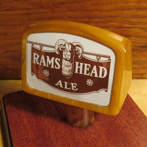 May include: A wooden beer tap handle with a white label that says "RAMS HEAD ALE" and features a brown illustration of a ram's head.
