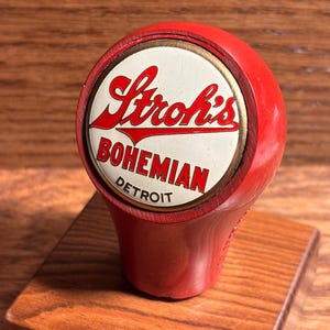 May include: A red Stroh's Bohemian Detroit tap handle. The handle has a white and red logo with the words "Stroh's Bohemian Detroit". The tap handle is mounted on a small wooden base.