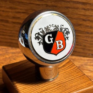 May include: A chrome gear shift knob with a white emblem featuring the letters "G" and "B" in black and red, surrounded by decorative flourishes. The knob is mounted on a square, wooden base with a polished finish.