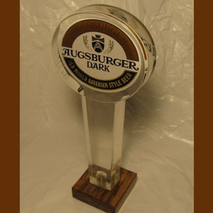 May include: A clear acrylic beer tap handle with a round, brown and white label that reads "Augsburger Dark Old World Bavarian Style Beer".