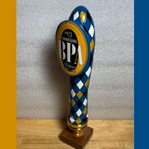 May include: A blue and gold beer tap handle with a diamond pattern and the text "OMMEGANG BPA". The tap handle has a wooden base and a gold-colored metal accent.