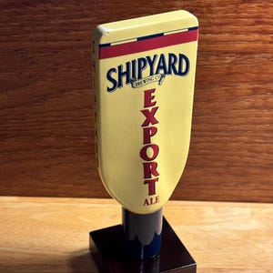 May include: A Shipyard Brewing Co. Export Ale tap handle. The handle is yellow with red and blue accents and the words "SHIPYARD BREWING CO. EXPORT ALE" printed on it. It sits on a dark brown square base.