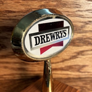 May include: A gold-colored metal bar top sign with a white oval featuring the text "DREWRYS" in black lettering and a red ribbon. The sign is mounted on a wooden base.