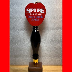 May include: A black and red draft cider tap handle with the text "SPIRE MOUNTAIN DRAFT CIDER APPLE". The handle has a red apple-shaped top and a wooden base. The handle is designed for dispensing draft cider.