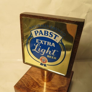 May include: A wooden bar tap handle with a gold-colored metal plate featuring the Pabst Blue Ribbon logo and the text "Naturally Brewed Pabst Extra Light Beer".