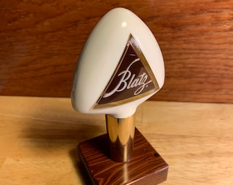 Könnte beinhalten: Ein weißer Keramik-Bierzapfhahn mit goldener Verzierung und einem braunen Logo mit der Aufschrift "Blatz". Der Hahn ist auf einem dunklen Holzsockel montiert.
