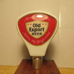 May include: A white beer tap handle with a red and white label that says "Mountain Water Old Export Beer Makes the Difference". The label has three stars on each side of the text "Old Export". The tap handle is on a dark brown wooden base.