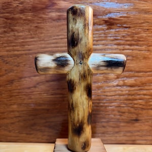May include: A handcrafted wooden cross with a natural finish, mounted on a small wooden base. The cross features a rustic design with dark brown and tan tones, showcasing the wood grain. The cross is a symbol of faith and spirituality.
