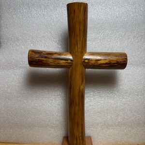 May include: A handcrafted wooden cross with a vertical post and two horizontal arms. The cross is made of light brown wood with darker brown grain patterns. It stands on a small, square wooden base.