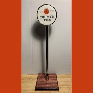 May include: A silver beer tap handle with a round, white sign that says "Trumer Pils" in black lettering. The sign has a red and gold logo in the center. The handle is mounted on a dark wood base.