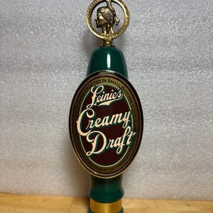 May include: A green and gold beer tap handle with the text "Leinie's Creamy Draft". The tap handle has a gold ring with a Native American profile. The text "Hand-Crafted in Small Batches" is also visible.