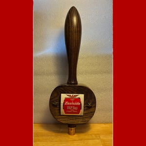 May include: A wooden beer tap handle with a dark brown finish. The handle has a rounded top and a circular base with a label that reads "Eastside Old Tap Lager Beer". Two small star-shaped accents flank the label.