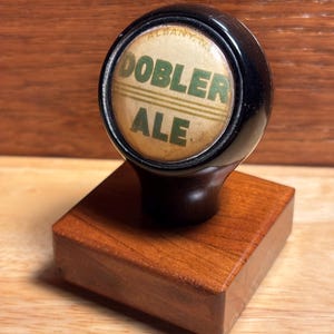May include: A vintage Dobler Ale tap handle. The handle has a black, rounded top with the words "DOBLER ALE" in green lettering. The handle is mounted on a square, wooden base with a rich, reddish-brown finish.