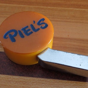 May include: A yellow and silver bottle opener with the word "PIEL'S" in blue lettering on the top.