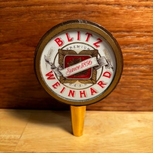 May include: A vintage Blitz-Weinhard beer tap handle with a gold-colored metal base and a round wooden top. The top features a white circular label with red and gold lettering that reads "Blitz-Weinhard Premium Beer Since 1856".
