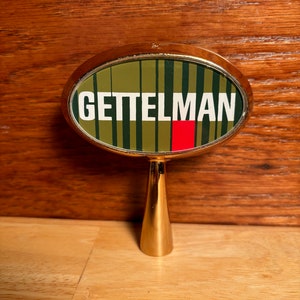 May include: A gold-colored metal tap handle with a round, green and black striped top. The top has a white oval with the word "GETTELMAN" in black letters and a red rectangle.