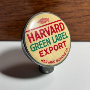 May include: A vintage beer tap handle with a circular face. The face is cream-colored with the words "HARVARD GREEN LABEL EXPORT" in red and green. The words "HARVARD BREWING CO." are also present. The handle is metal.