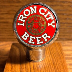 May include: A round metal beer tap handle featuring a red circular center with the white text IRON CITY BEER. In the middle, a smaller circular emblem displays the text PITTSBURGH BREWING CO and a red graphic of a stylized animal head.