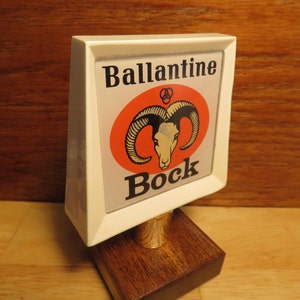 May include: White plastic beer tap handle with a red, white, and black logo. The logo features a ram's head and the text "Ballantine Bock".