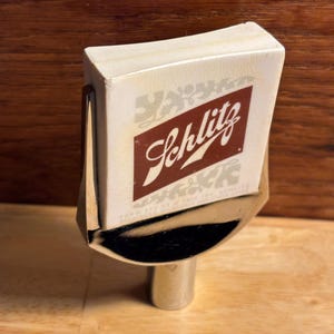 May include: A vintage Schlitz beer tap handle. The handle features a rectangular white top with the Schlitz logo in a maroon diamond. The tap is made of chrome and has a curved design.