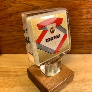 May include: A vintage Duke Beer tap handle. The tap handle has a clear, rectangular body with a gold, red, and black design. The base is a square of stained wood. The tap handle is designed to be mounted on a beer tap.