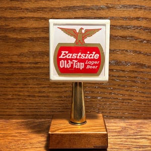 May include: A vintage beer tap handle with a white rectangular top featuring the words "Eastside Old Tap Lager Beer" in red and gold. An eagle graphic is above the text. The handle has a gold-colored stem and a wooden base.